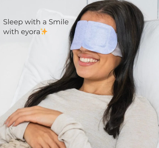 Woman wearing a sleep mask with 'eyora' branding, smiling and lying on a white pillow.